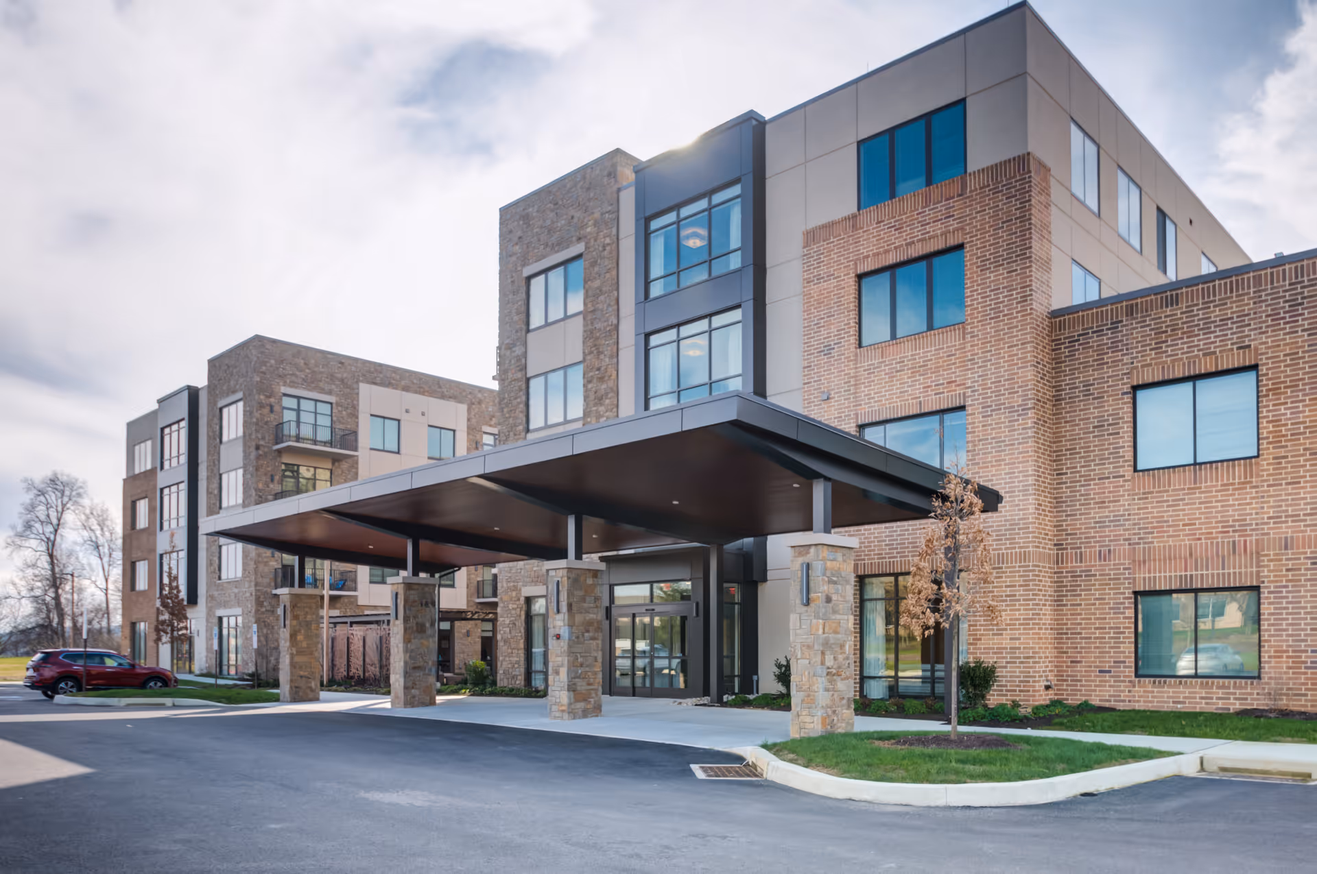 Exterior view of a modern multi-story senior living facility with a covered entrance supported by stone pillars, large windows, and a mix of brick and stone facade under a partly cloudy sky.