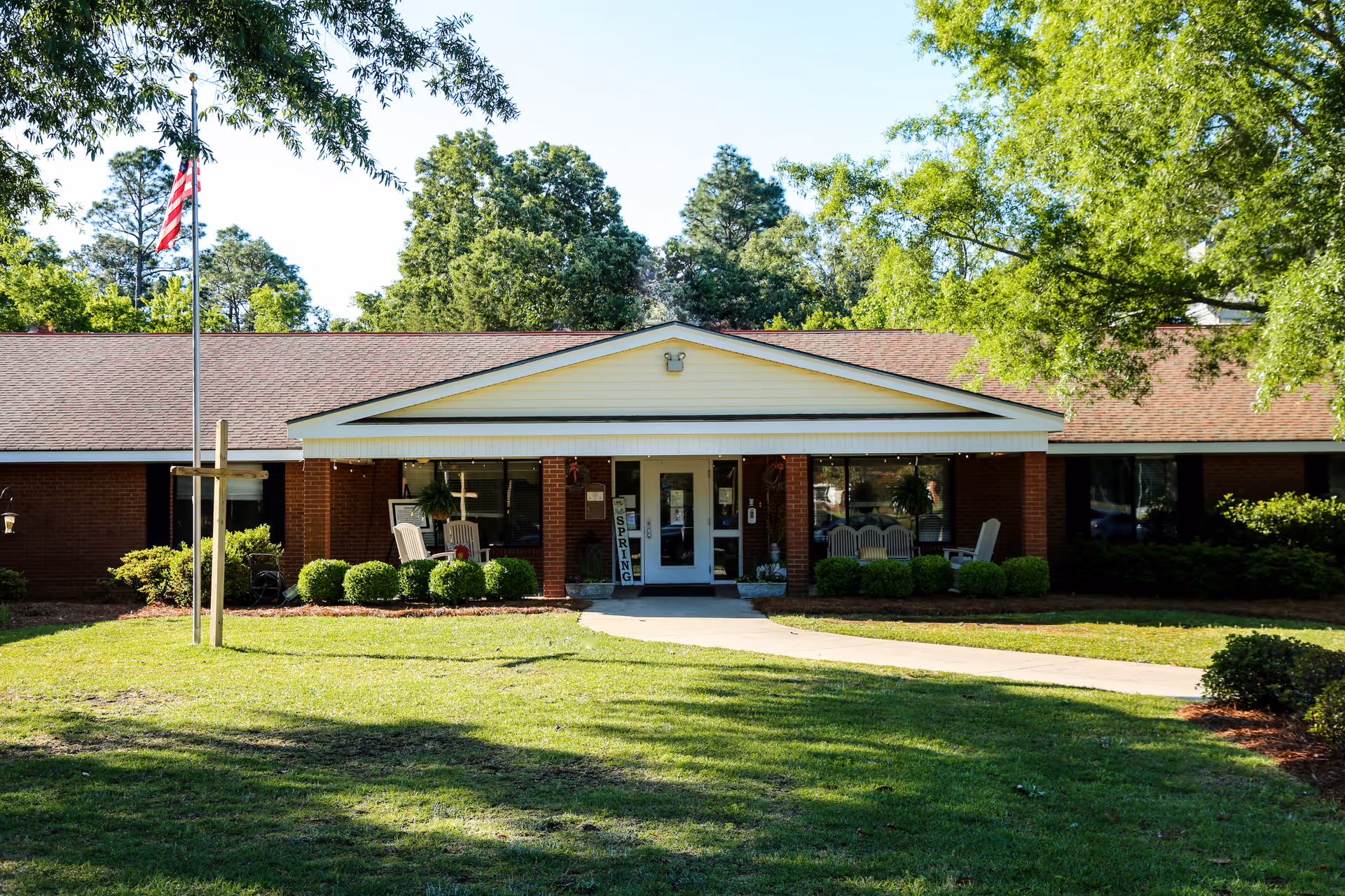 Front exterior view of a single-story brick building with a peaked roof and a covered entrance. There are several chairs on the porch, neatly trimmed bushes, a flagpole with an American flag, and a well-maintained lawn with trees in the background.