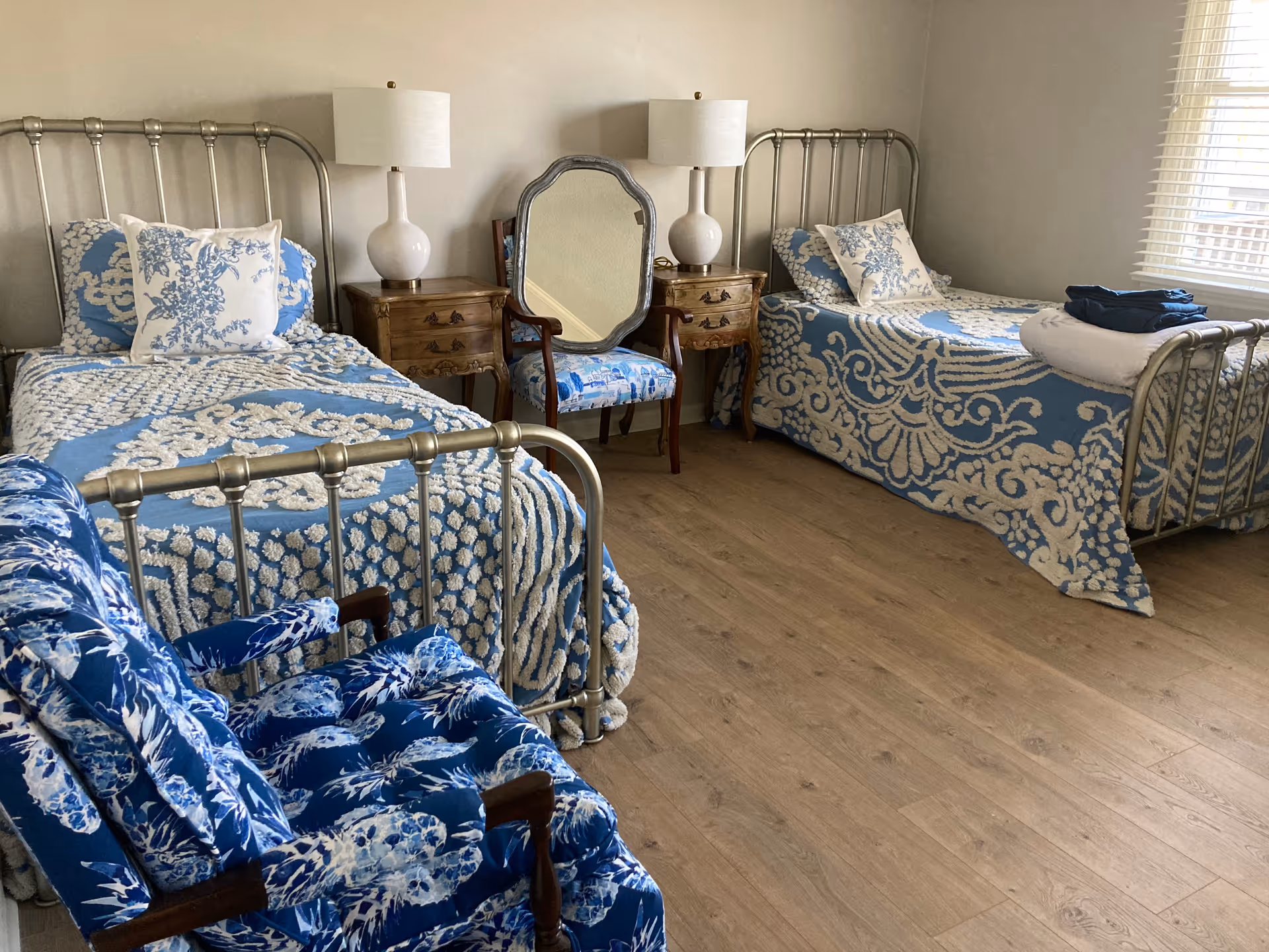 A bedroom with two twin beds featuring metal frames and blue and white patterned bedding. Between the beds are two wooden nightstands with white lamps. A wooden chair with blue and white upholstery and a large mirror behind it are positioned between the nightstands. The room has light-colored walls and a window with blinds on the right side.