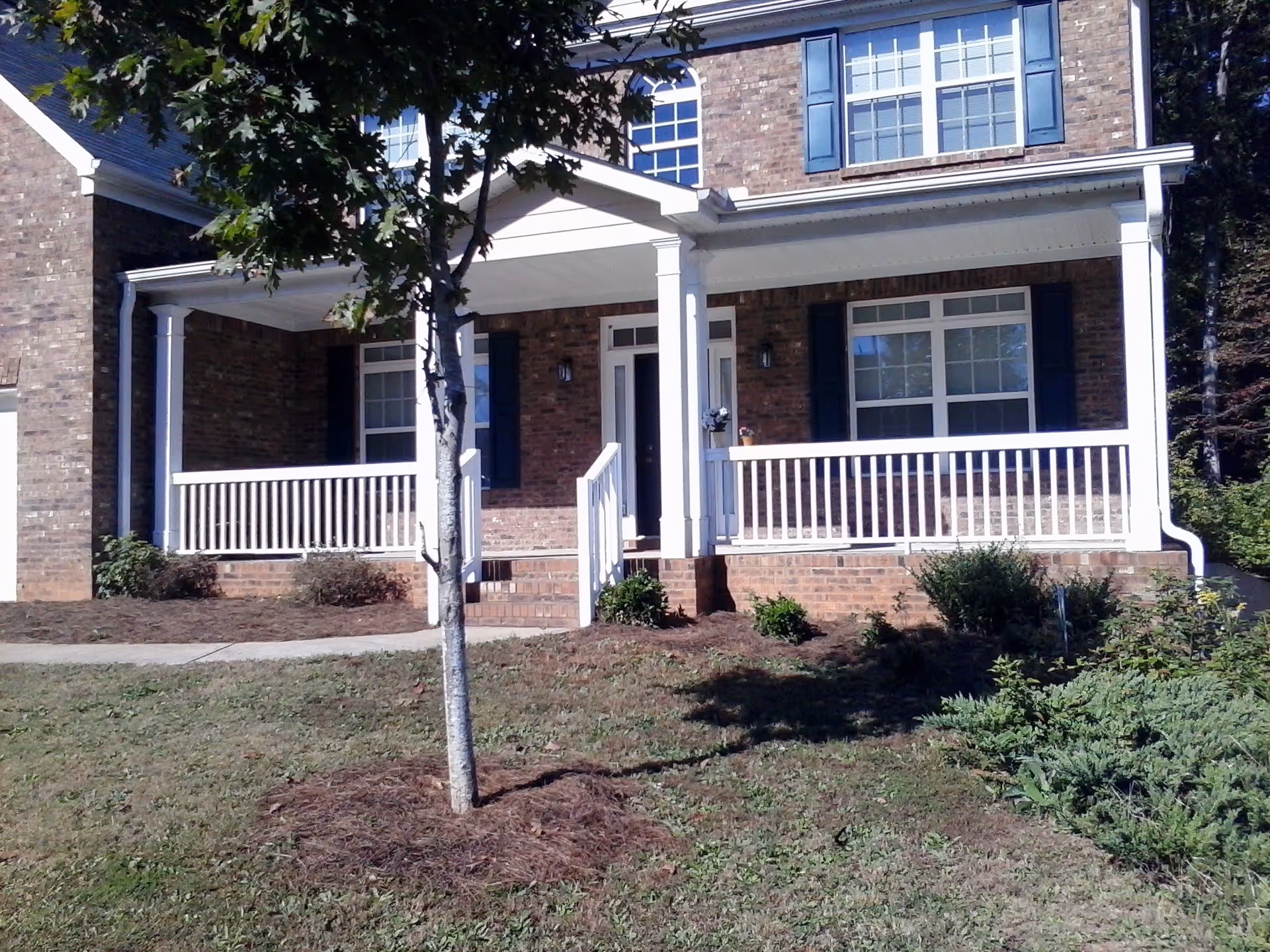 Front exterior view of a brick house with a covered porch, white railings, and steps leading up to the front door. There is a tree and some shrubs in the front yard.