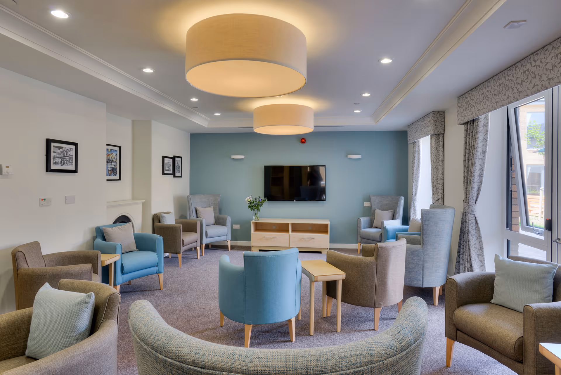 A spacious and well-lit living room with multiple armchairs and small tables arranged in a circle facing a wall-mounted flat-screen TV. The room has large windows with patterned curtains, light blue and beige walls, and two large round ceiling lights. There are framed pictures on one wall and a small cabinet with a vase of flowers beneath the TV.