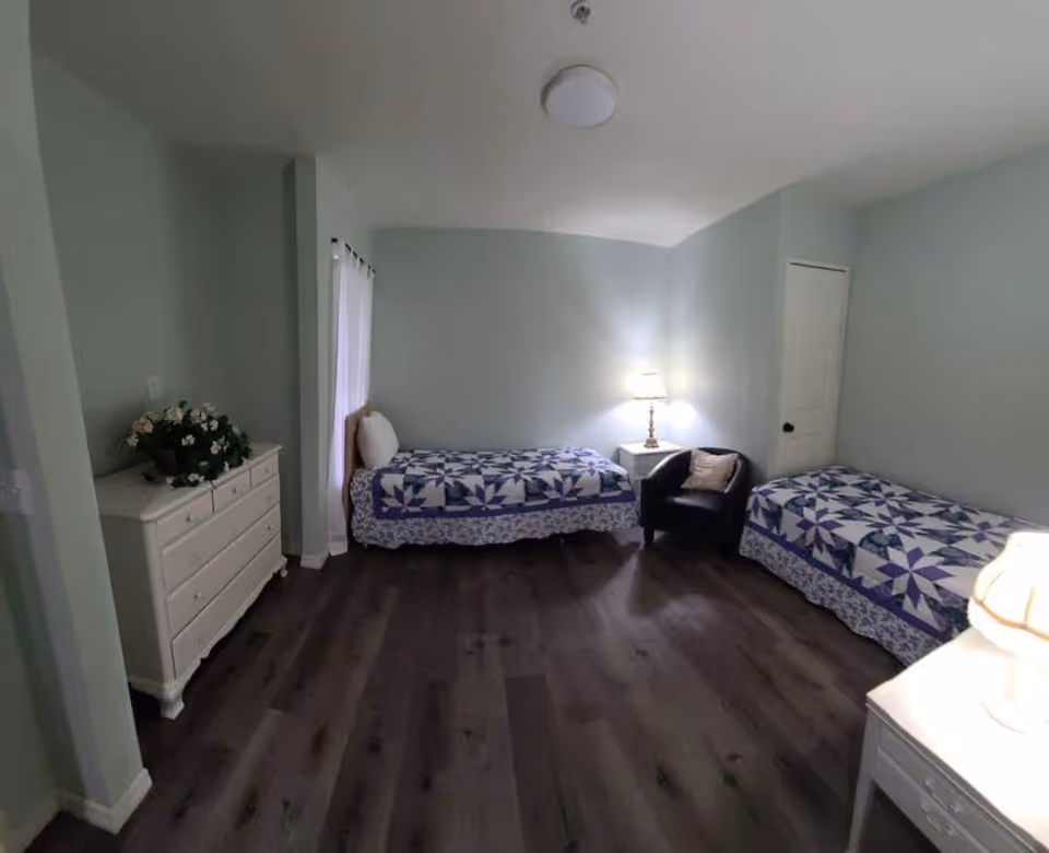 A bedroom with two twin beds covered with matching blue and white patterned quilts. There is a white dresser with a flower arrangement on top, a small white nightstand with a lamp, and a black armchair with a pillow. The walls are painted light blue and the floor is dark wood.