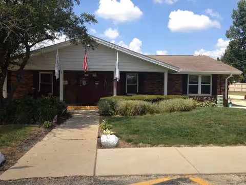 Front exterior of a single-story senior living center with flags, shrubs, and walkway.