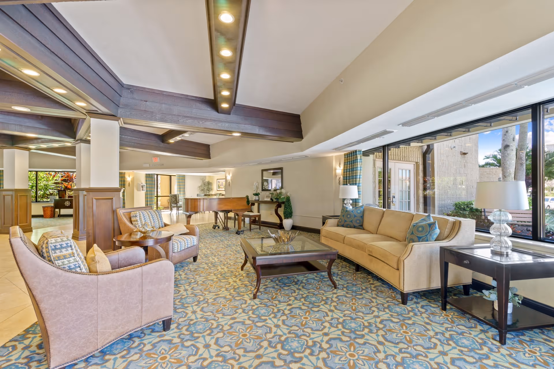 A spacious and well-lit living room area in a senior living facility featuring a beige sofa with blue patterned pillows, two armchairs with patterned cushions, a glass-top coffee table, and side tables with lamps. The room has large windows allowing natural light to enter, a patterned carpet with blue and beige tones, and a grand piano in the background near a console table with decorative plants and a mirror above it.