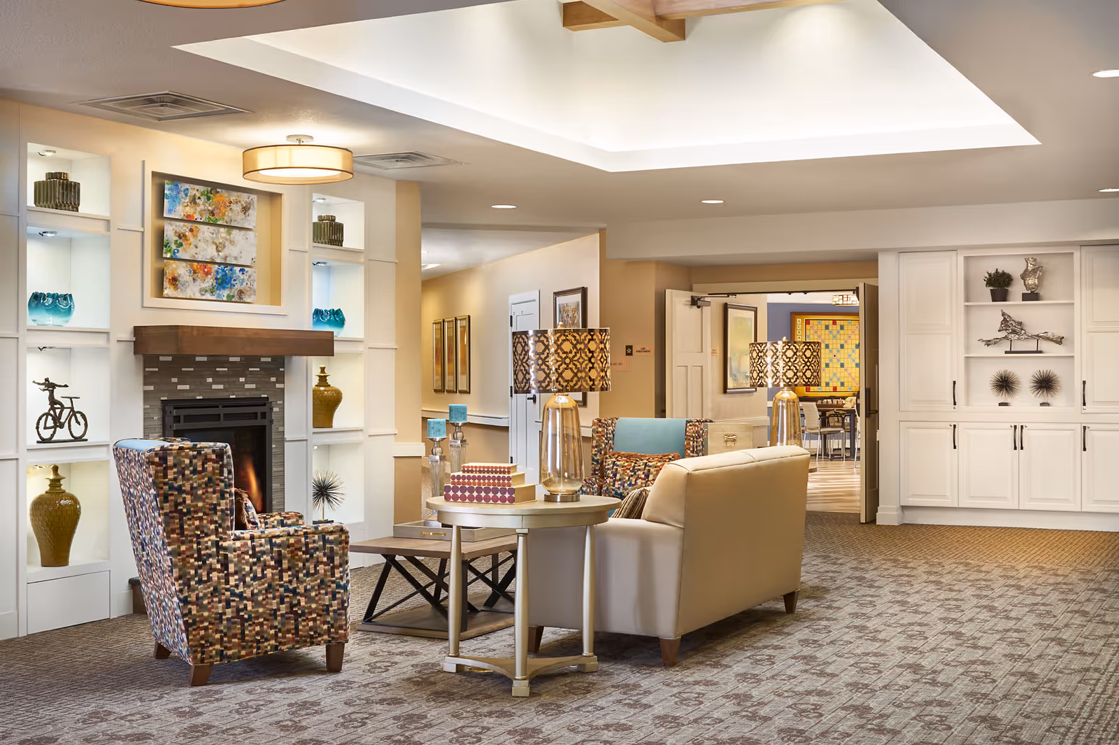 A cozy and well-lit living room area in an assisted living facility featuring patterned armchairs, a beige sofa, a round side table with decorative lamps, and a fireplace with built-in shelves displaying vases and art pieces. The room has a carpeted floor and a recessed ceiling light.