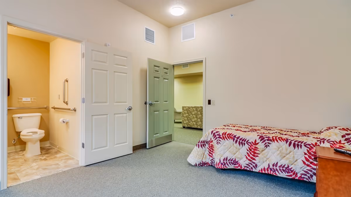 Assisted-living bedroom with a single bed covered in a red-and-white leaf-pattern quilt, open doors showing a grab-bar equipped bathroom and a small sitting area.