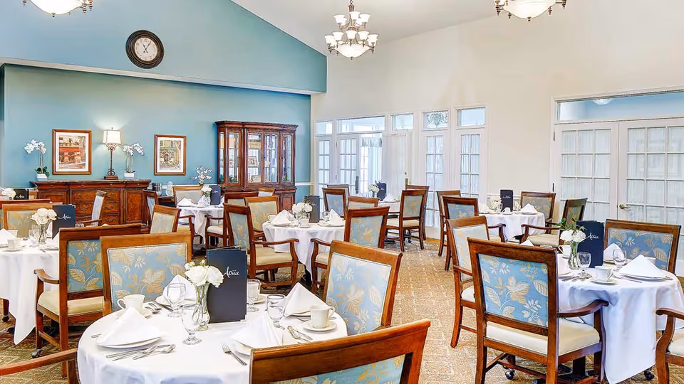 A bright and elegant dining room with multiple round tables covered in white tablecloths, each set with white napkins, cups, glasses, and silverware. The chairs have wooden frames with blue floral upholstery. The room features a blue accent wall with framed artwork, a wooden cabinet, and a clock above. Large windows and glass doors allow natural light to fill the space.