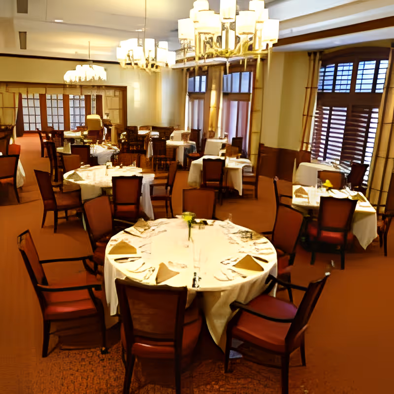 A spacious dining room with multiple round tables covered in white tablecloths, each set with folded napkins and silverware. The room features wooden chairs with red cushions, large windows with wooden shutters, and elegant chandeliers hanging from the ceiling.