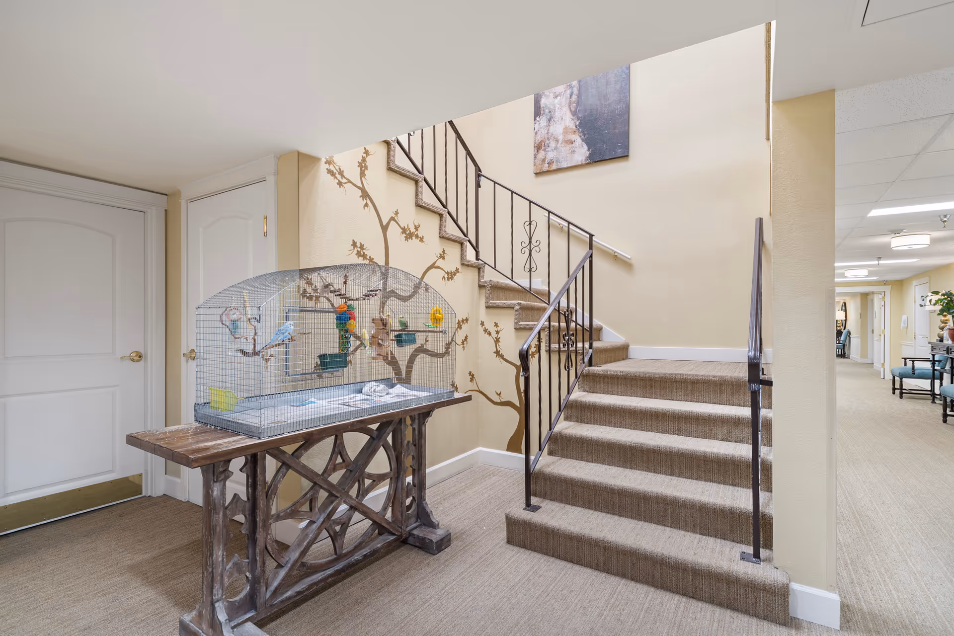 Interior common area with a carpeted staircase, wrought-iron railing, a decorative table holding a large birdcage, and a hallway leading to seating.