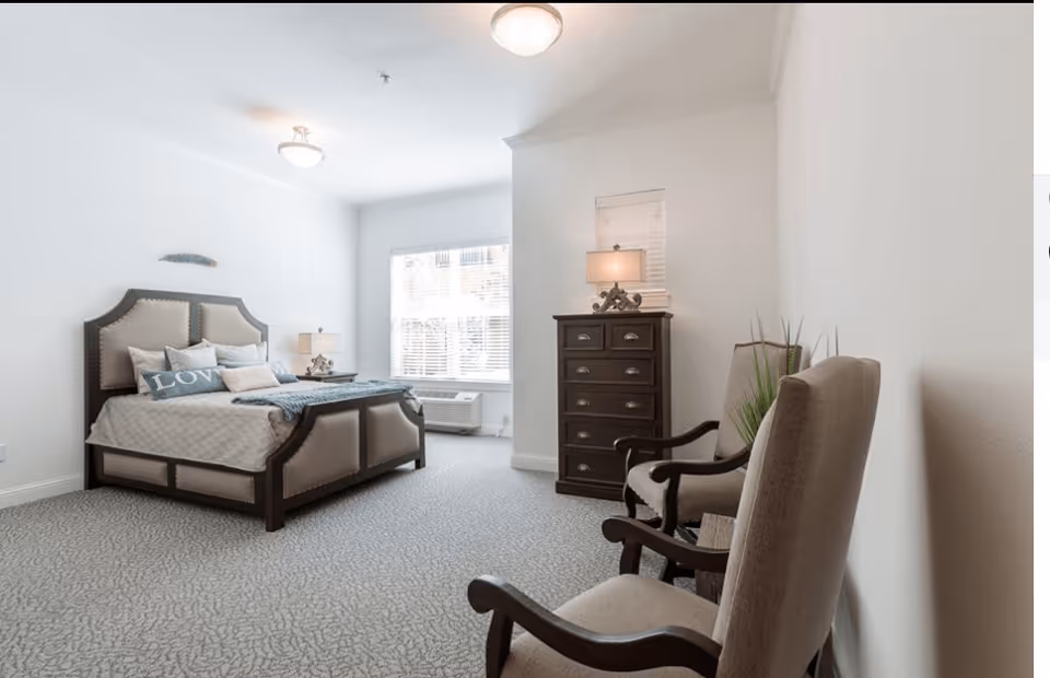 Bright, neutral bedroom with an upholstered bed, bedside table, tall dresser, and two armchairs facing a window.