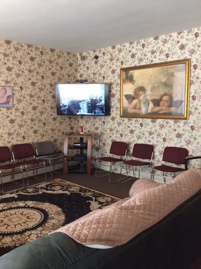 A cozy living room with floral wallpaper, a patterned area rug, a row of maroon and gray chairs along the wall, a flat-screen TV mounted on the wall above a small wooden stand with electronic devices, and a framed painting of two cherubs labeled 'ANGELS'. A green couch with a pink quilted cover is partially visible in the foreground.