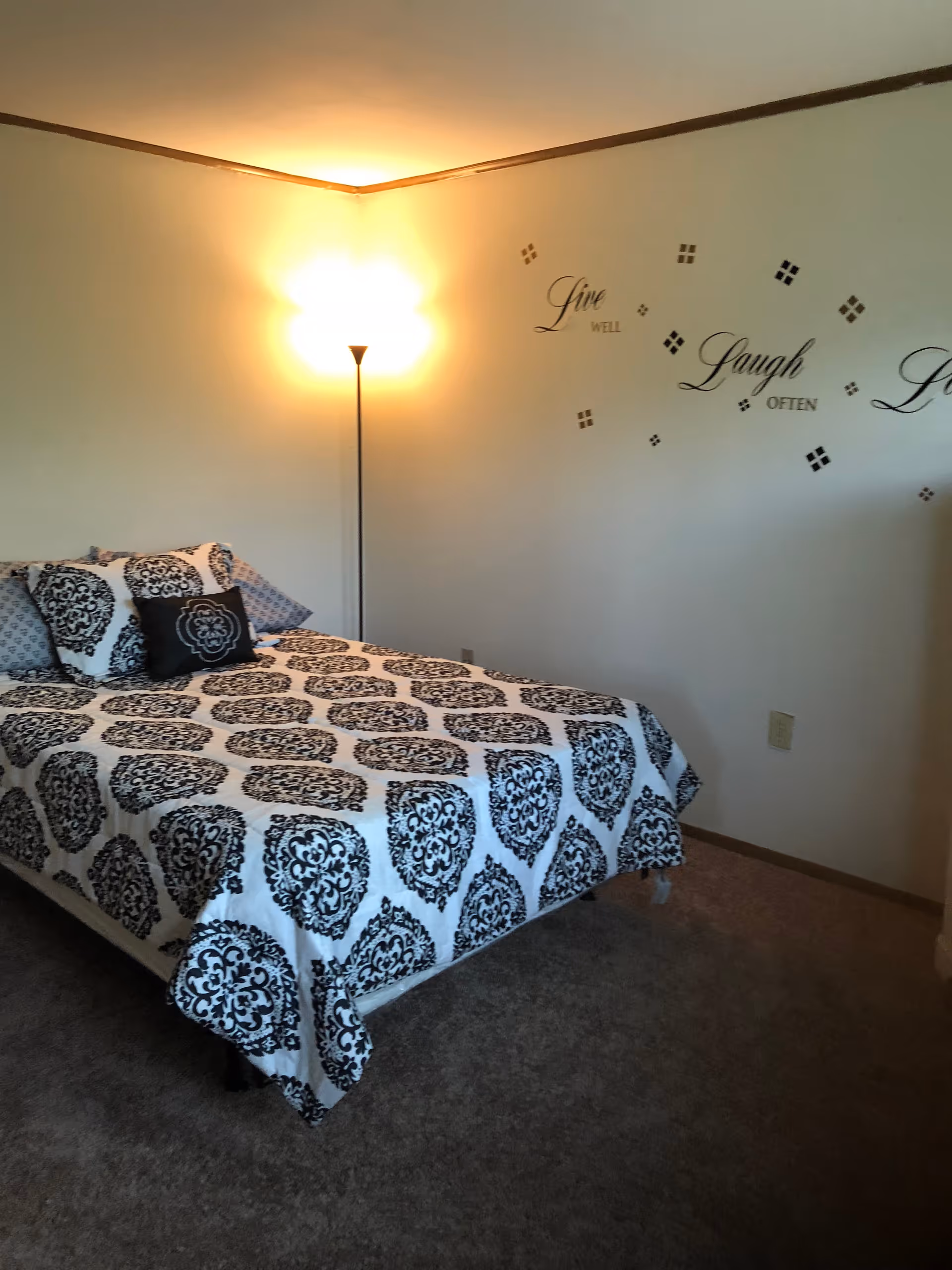 A bedroom with a bed covered in a black and white patterned bedspread and matching pillows. A floor lamp is turned on in the corner, casting light on the wall. The wall has decorative text that reads 'Live well Laugh often'. The room has carpeted flooring and simple decor.