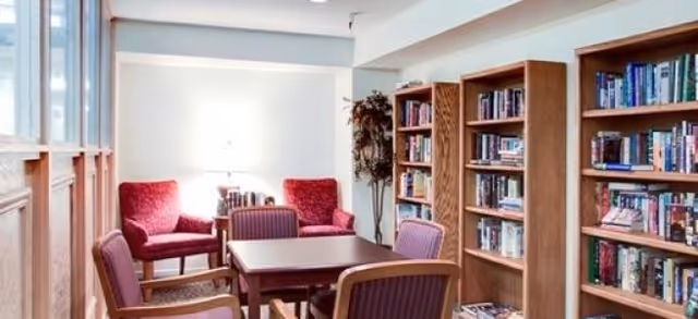A cozy reading and sitting area with wooden bookshelves filled with books, a square wooden table surrounded by four purple cushioned chairs, and two red armchairs near a window letting in natural light. A tall plant is placed in the corner of the room.