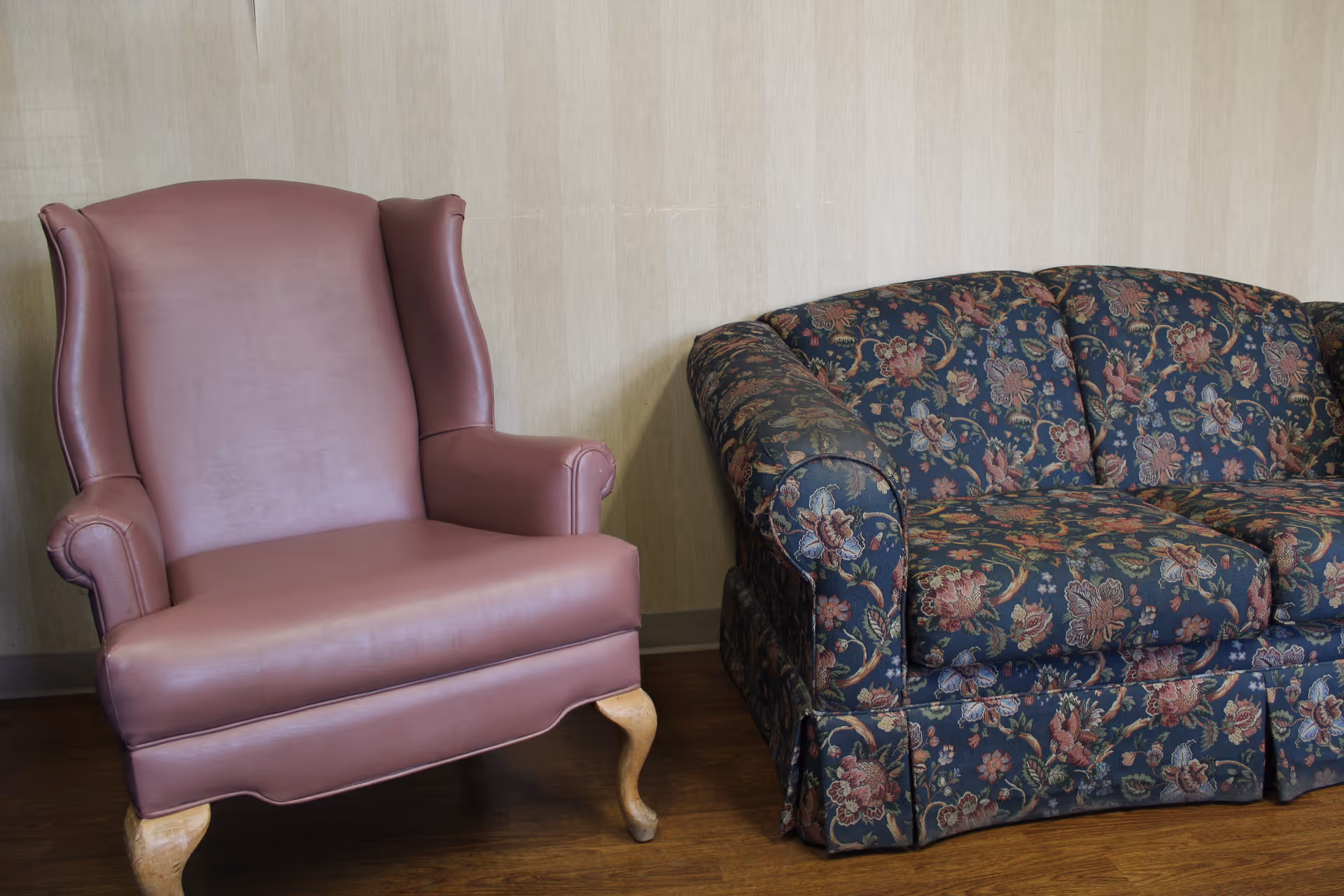 A maroon leather armchair next to a dark floral upholstered sofa against a beige wall on a wooden floor.