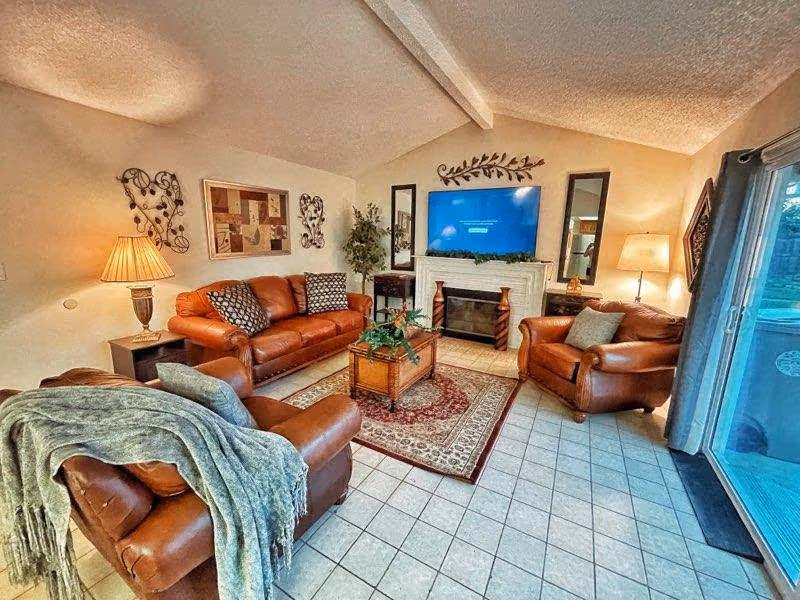 Cozy living room with leather sofas and armchairs arranged around a coffee table and rug, a TV above a fireplace, lamps, and sliding glass doors.