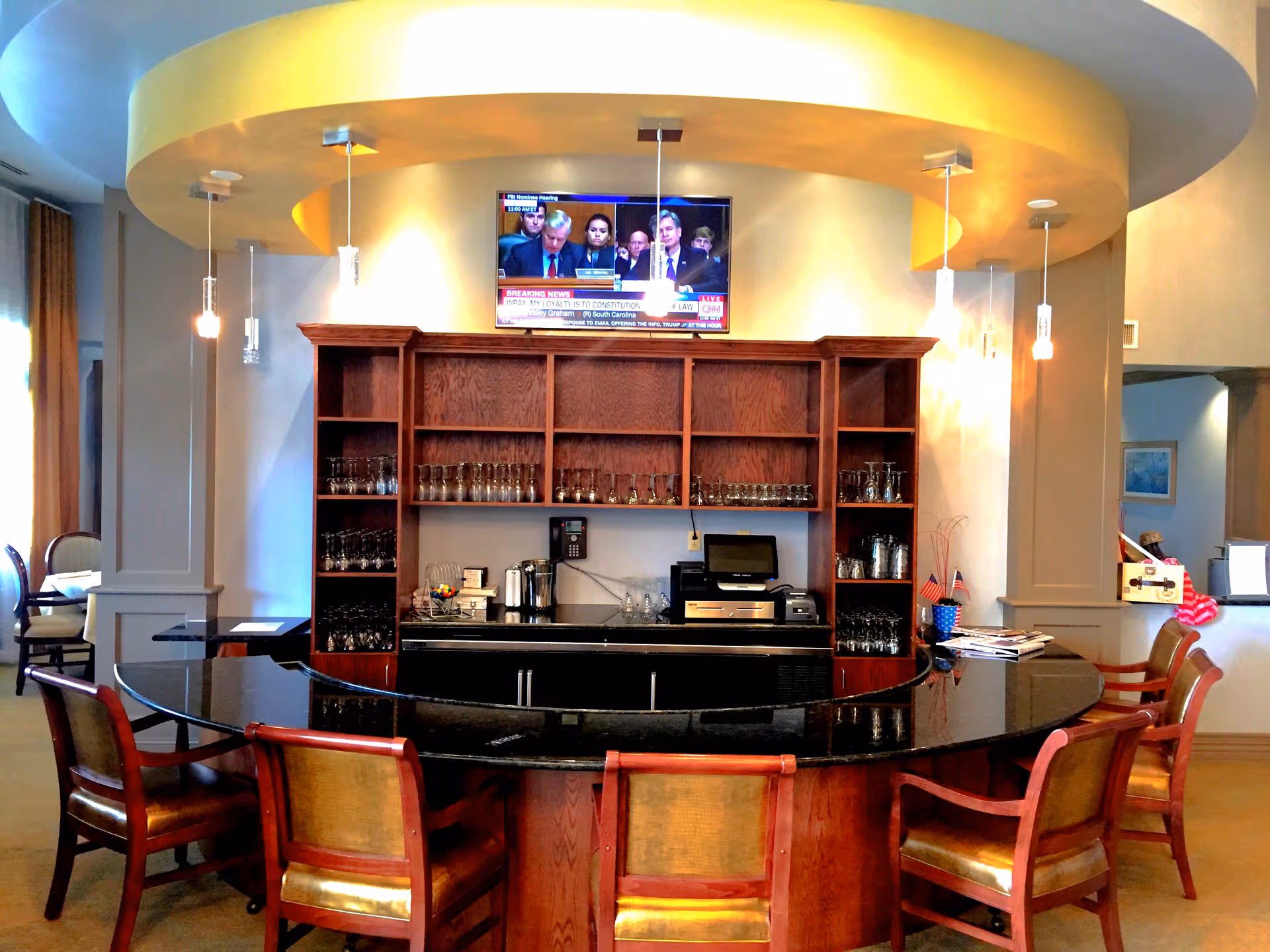 Curved community bar with high-backed chairs in front of wooden shelving stocked with glassware and a TV mounted above.