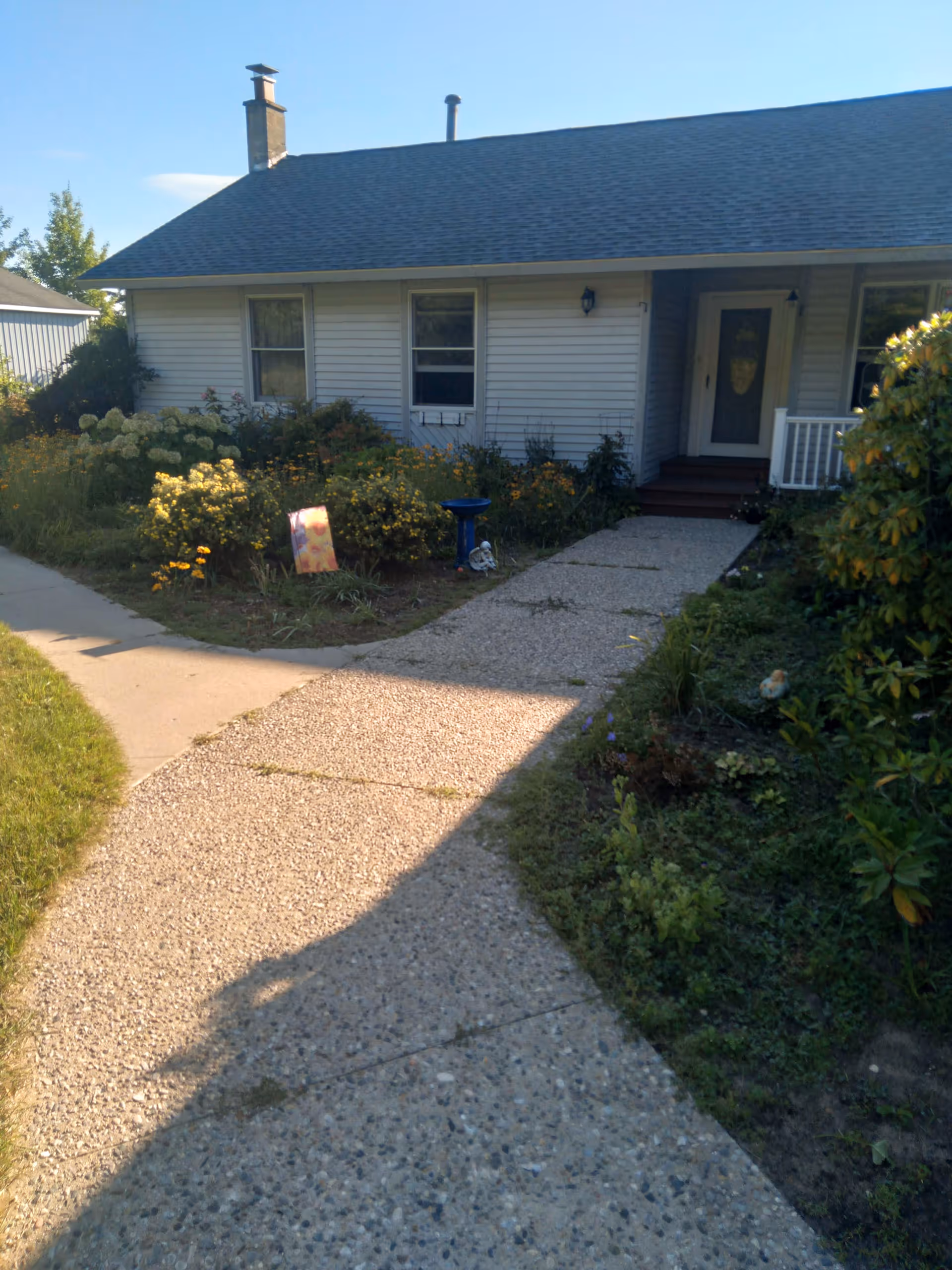 Front exterior view of a single-story house with a gray roof and white siding. There is a concrete pathway leading to the front door, surrounded by green bushes and flowering plants on both sides. The house has two windows visible and a small porch with steps.