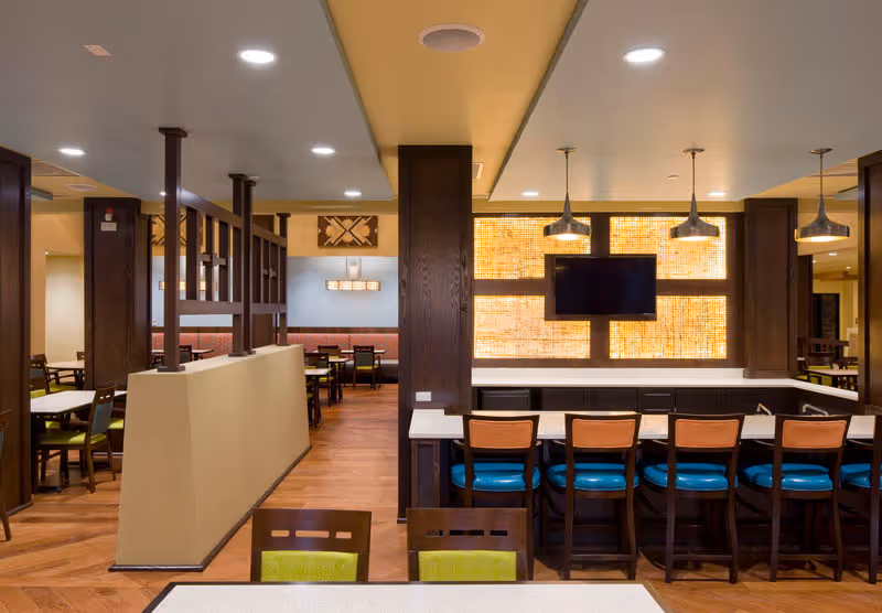 Interior view of a dining area in a senior living facility with wooden floors, multiple tables and chairs, a counter with high chairs, pendant lights, and a wall-mounted TV with a decorative illuminated panel behind it.