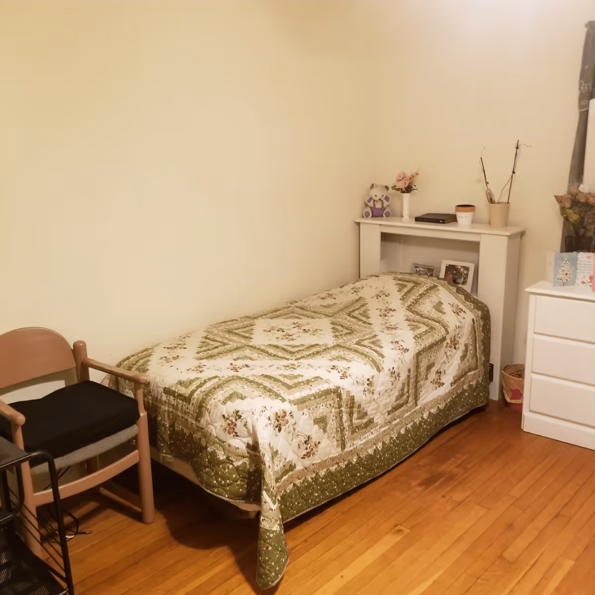 A small bedroom with a single bed covered in a green and white patterned quilt. Next to the bed is a white nightstand with a teddy bear, a vase with flowers, a small pot, and some framed photos. There is a wooden chair with a black cushion beside the bed, and a white dresser with greeting cards and flowers on top. The floor is wooden, and the walls are plain and light-colored.