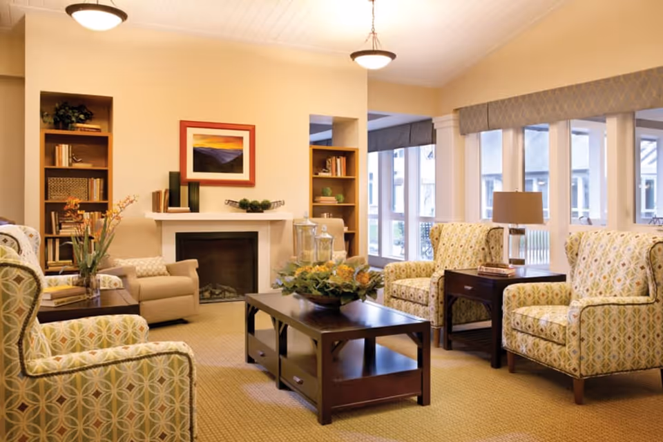 A cozy living room with patterned armchairs arranged around a dark wooden coffee table with a floral centerpiece. There is a fireplace with decorative items on the mantel and built-in bookshelves on either side. Large windows with valances allow natural light to fill the room.