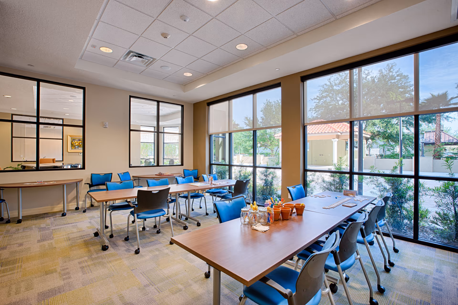 A bright activity room in a senior living facility with large windows letting in natural light. The room contains several tables with blue chairs arranged around them. On the tables, there are various craft supplies and games. The walls have large windows looking into another room, and outside greenery is visible through the exterior windows.
