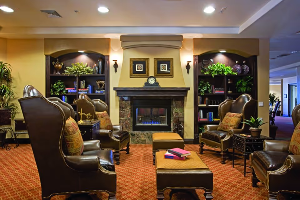 A cozy living room area with four brown leather armchairs arranged around two matching ottomans. The room features a fireplace with a clock and two framed pictures above it. On either side of the fireplace are built-in shelves decorated with plants, books, and decorative items. The floor is carpeted in a red patterned design, and the walls are painted a warm beige color.