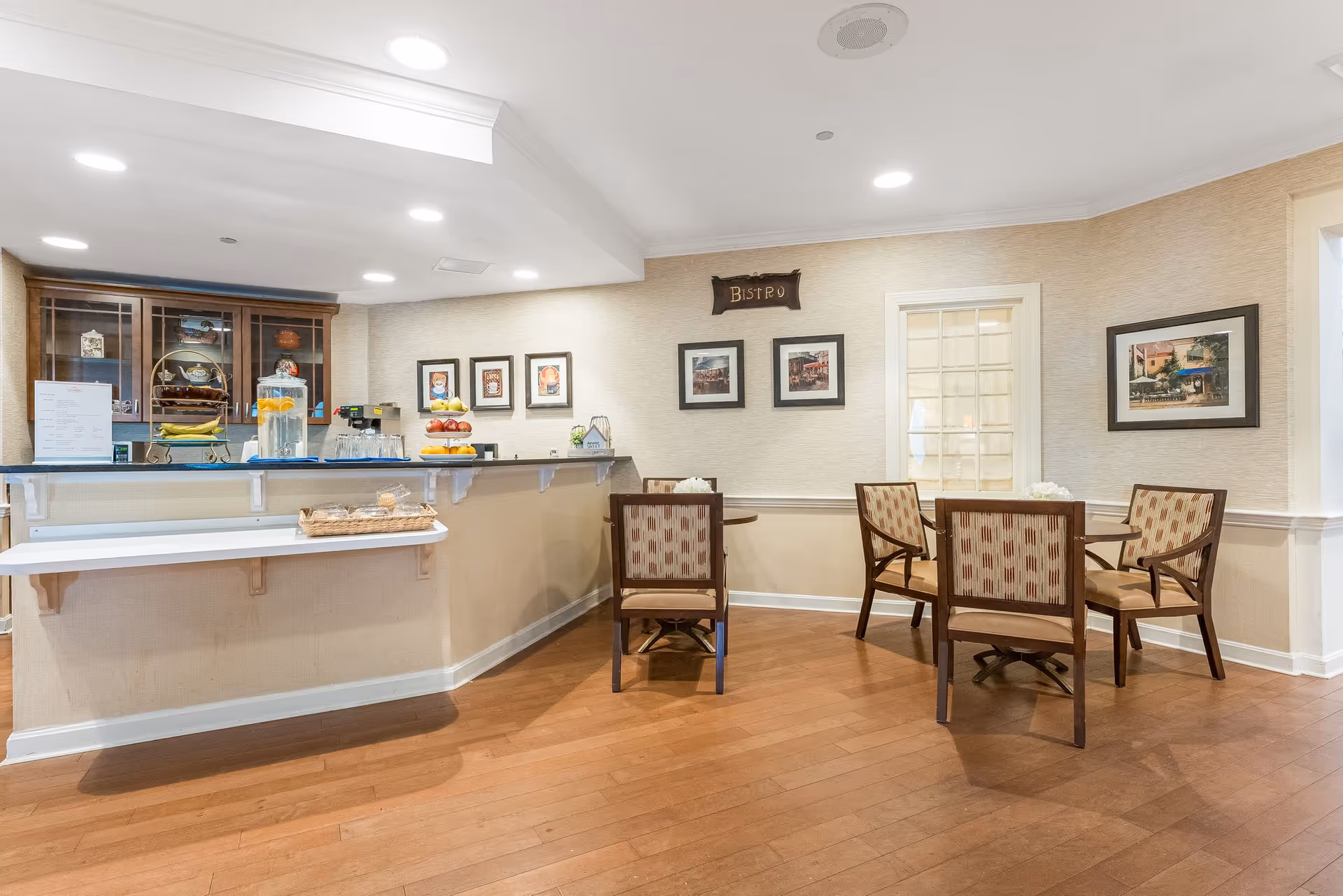 A cozy dining area in a senior living facility featuring a small round table with four cushioned chairs. The room has wooden flooring and beige walls adorned with framed pictures. A counter with a glass of water dispenser, fruit basket, and other items is visible on the left side under bright ceiling lights. A sign above the table reads 'Bistro'.