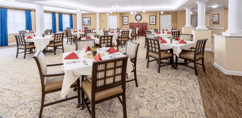 A spacious dining room with multiple round tables covered with white tablecloths, each set with red folded napkins, glassware, and silverware. The room features patterned carpet, beige walls, blue curtains on large windows, and decorative columns. There is a red sideboard with a clock and framed pictures on the far wall.