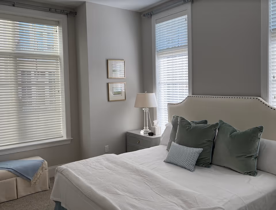 Bright bedroom featuring a bed with a padded headboard and green pillows, a nightstand with a lamp, and tall windows with blinds.