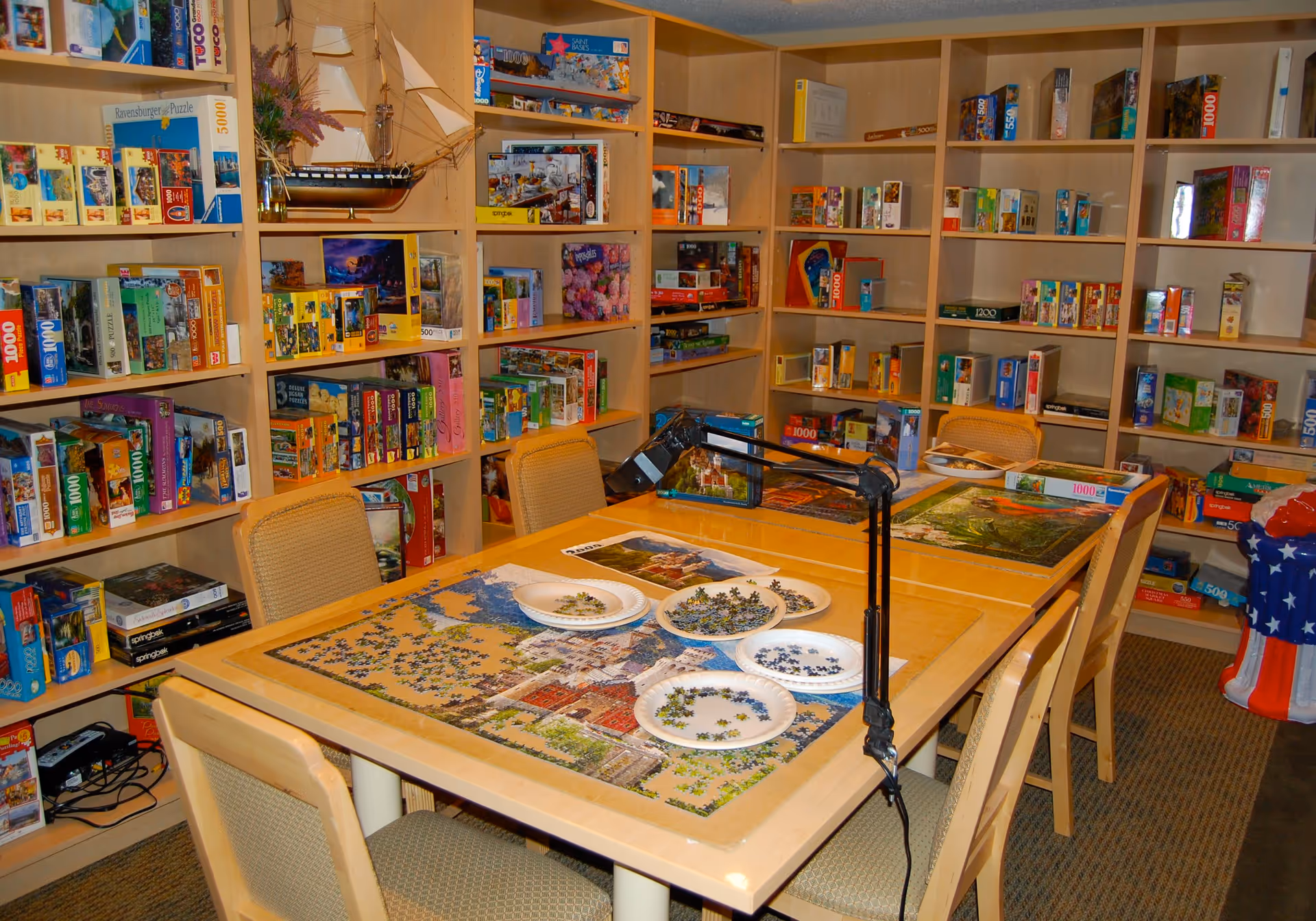 A room with shelves filled with numerous puzzle boxes and a table in the center where several puzzles are being assembled. The table has plates with puzzle pieces and a lamp attached to it. Four chairs surround the table.