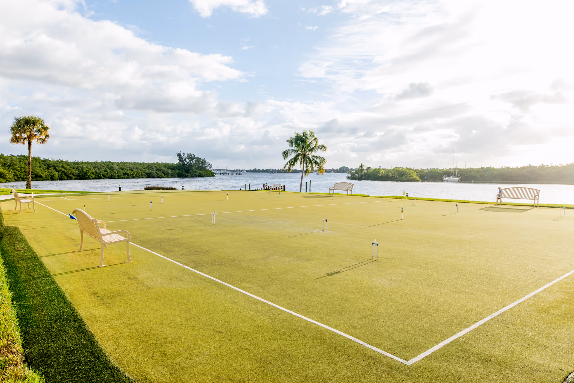 A waterfront lawn bowling green with benches and palm trees overlooking a river and a sailboat.