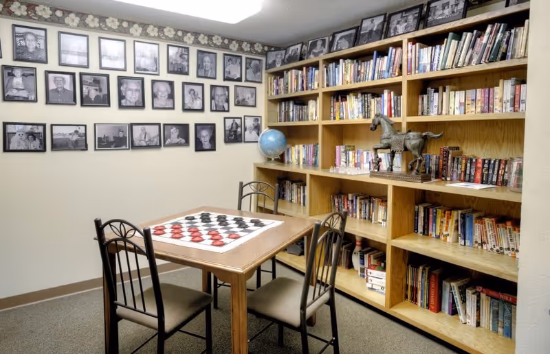 A cozy room with a wooden table and four chairs, featuring a checkers board on the table. One wall is decorated with framed black and white photographs, and the adjacent wall has large wooden bookshelves filled with books, a globe, and a horse statue.