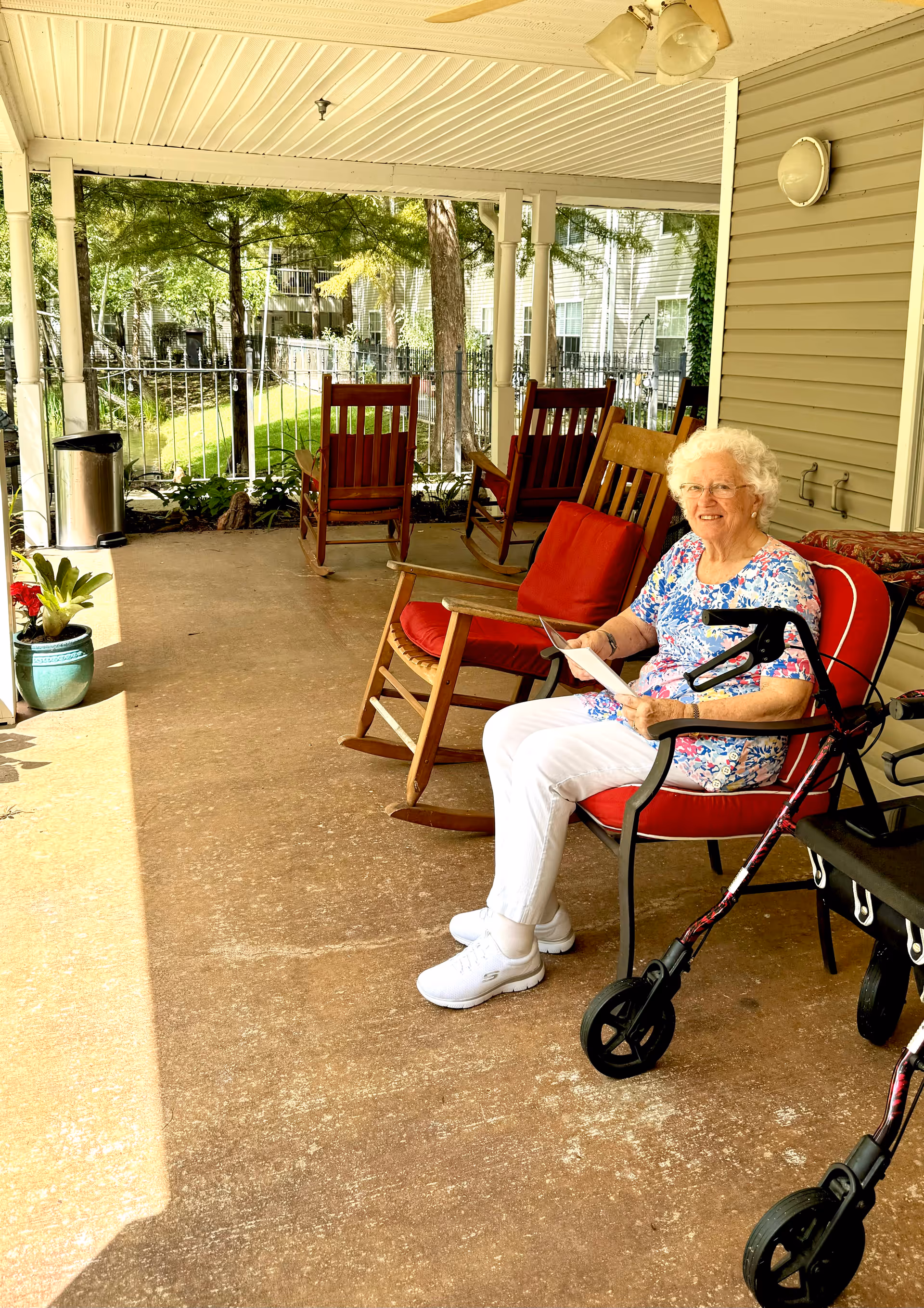 An elderly woman with white hair and glasses sits on a red cushioned chair on a covered patio. She is wearing a floral shirt and white pants, holding a piece of paper. Next to her is a walker with large wheels. The patio has several wooden rocking chairs with red cushions, potted plants, and overlooks a green outdoor area with trees and a fence.