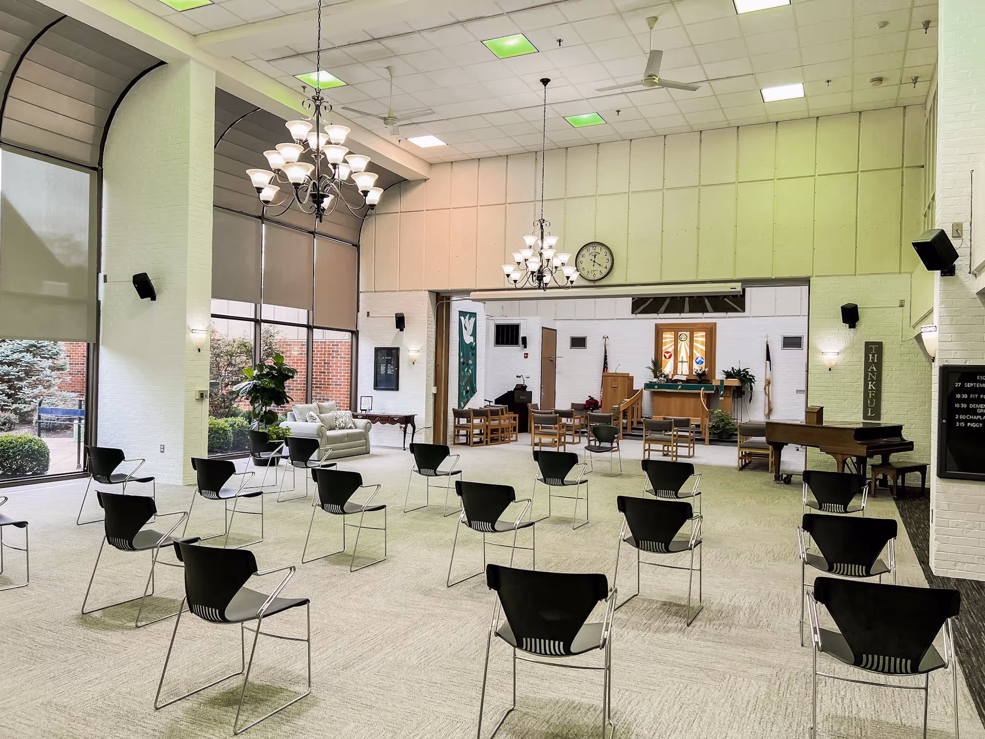 A spacious room with rows of black chairs arranged facing a small stage area with a podium, chairs, and a piano. The room has large windows with shades, two chandeliers hanging from the ceiling, and a clock on the wall above the stage. There are plants and a sofa near the windows, and banners and flags near the stage area.