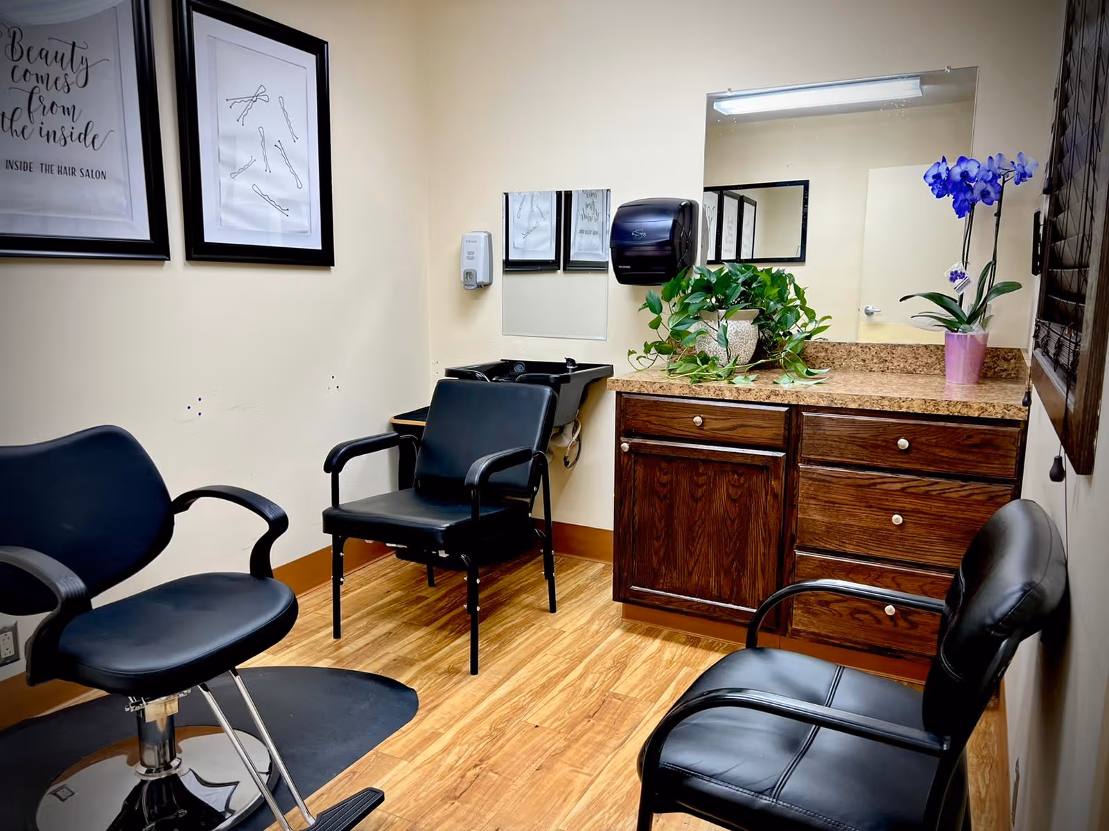 Small hair salon inside a care facility with styling chairs, a shampoo sink, mirror, wooden cabinets and potted plants.