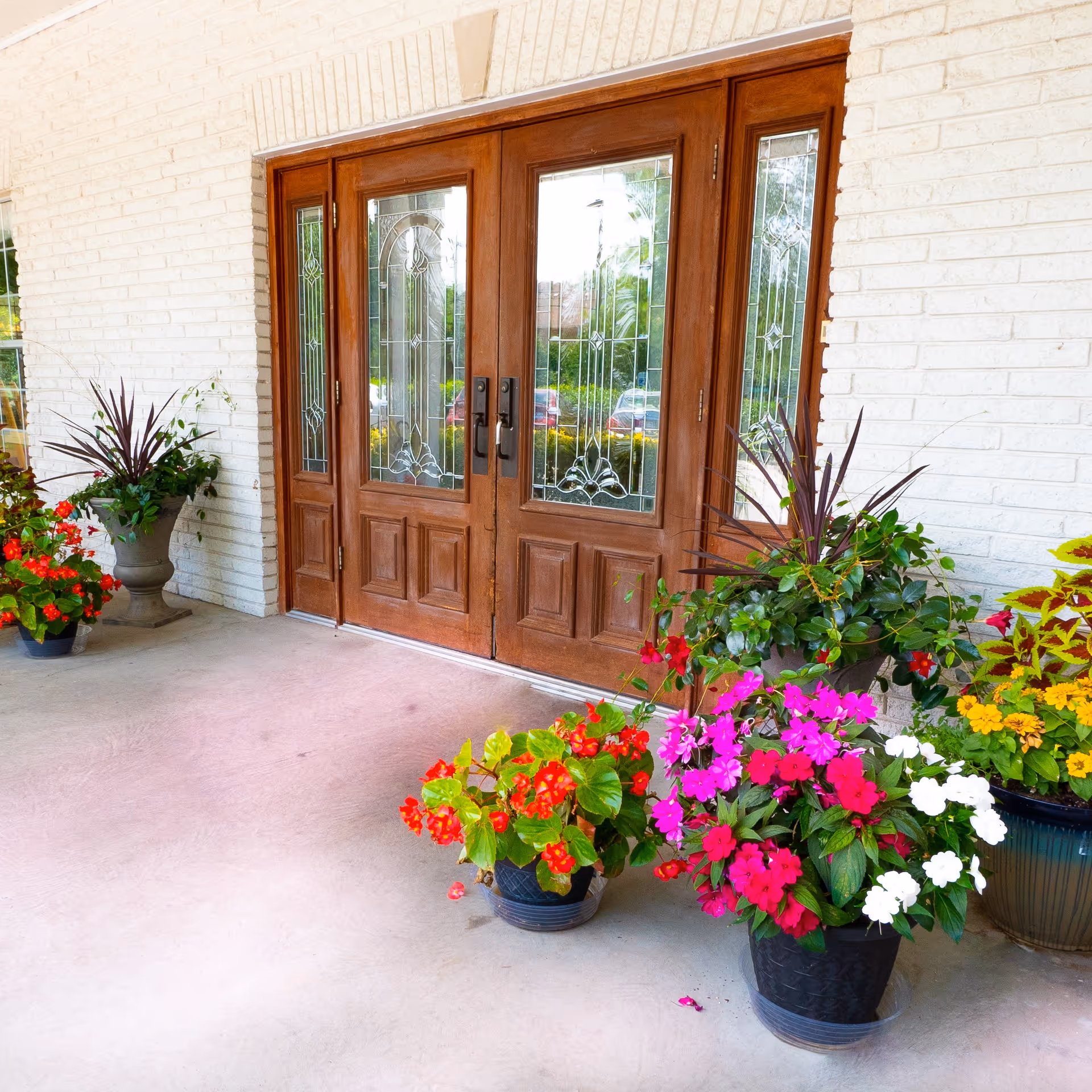 Wooden double front doors with decorative glass panels flanked by colorful potted flowers on a covered entrance porch.