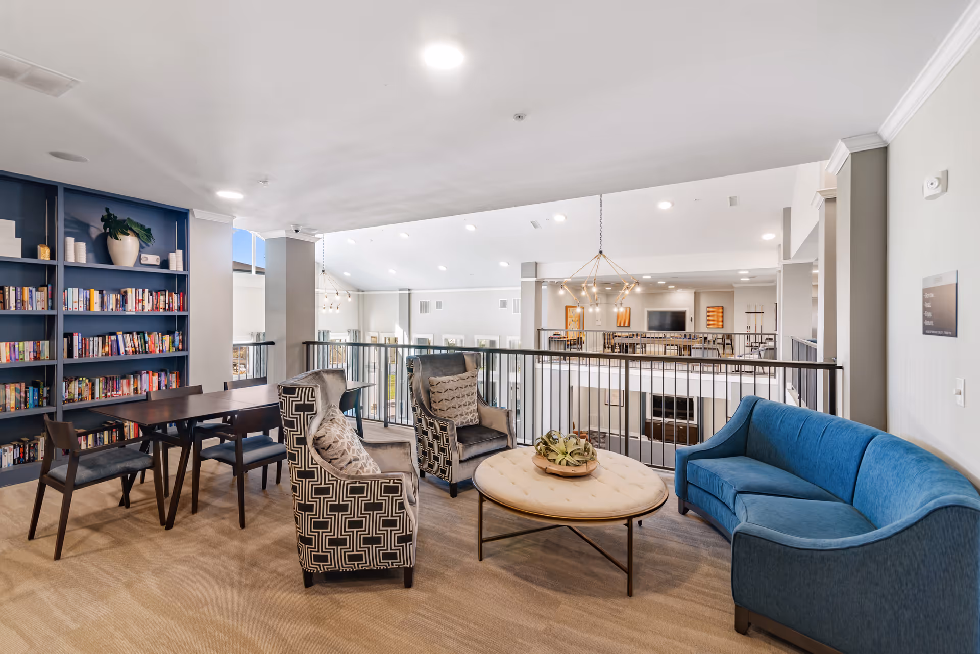 A bright and spacious lounge area with a blue couch, two patterned armchairs, and a round coffee table with a decorative centerpiece. Behind the seating area is a wooden table with four chairs and a large bookshelf filled with books. The space overlooks a lower level with additional seating and a fireplace, illuminated by modern chandeliers and recessed lighting.