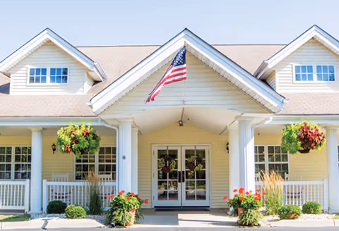 Front entrance of a light-colored building with columns, an American flag, hanging flower baskets, and double glass doors.