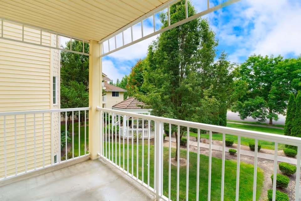View from a covered balcony with white railing overlooking landscaped grounds, trees, and a gazebo.