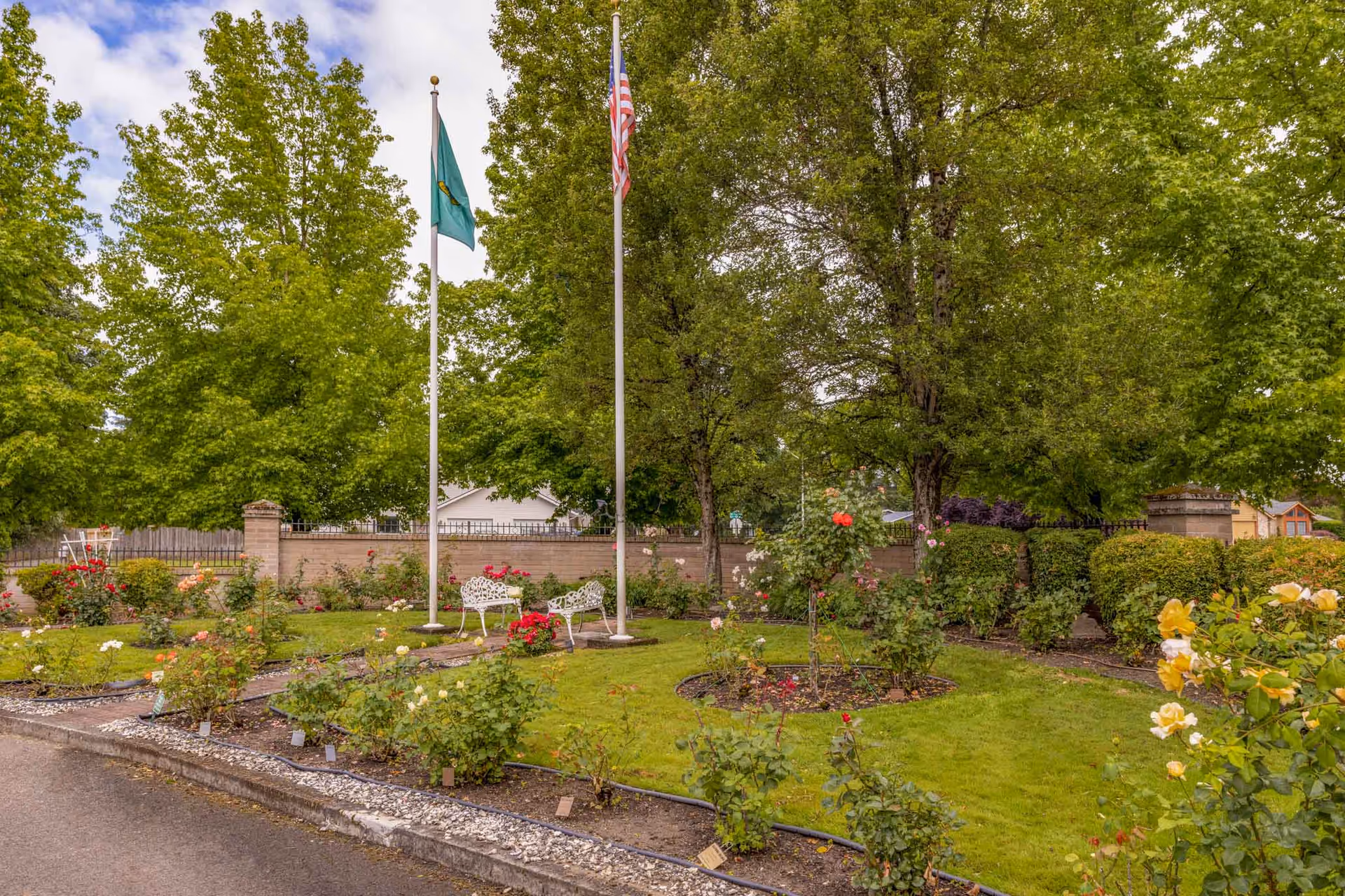 Landscaped outdoor garden with two flagpoles, white benches, rose beds, and large trees by a low brick wall.