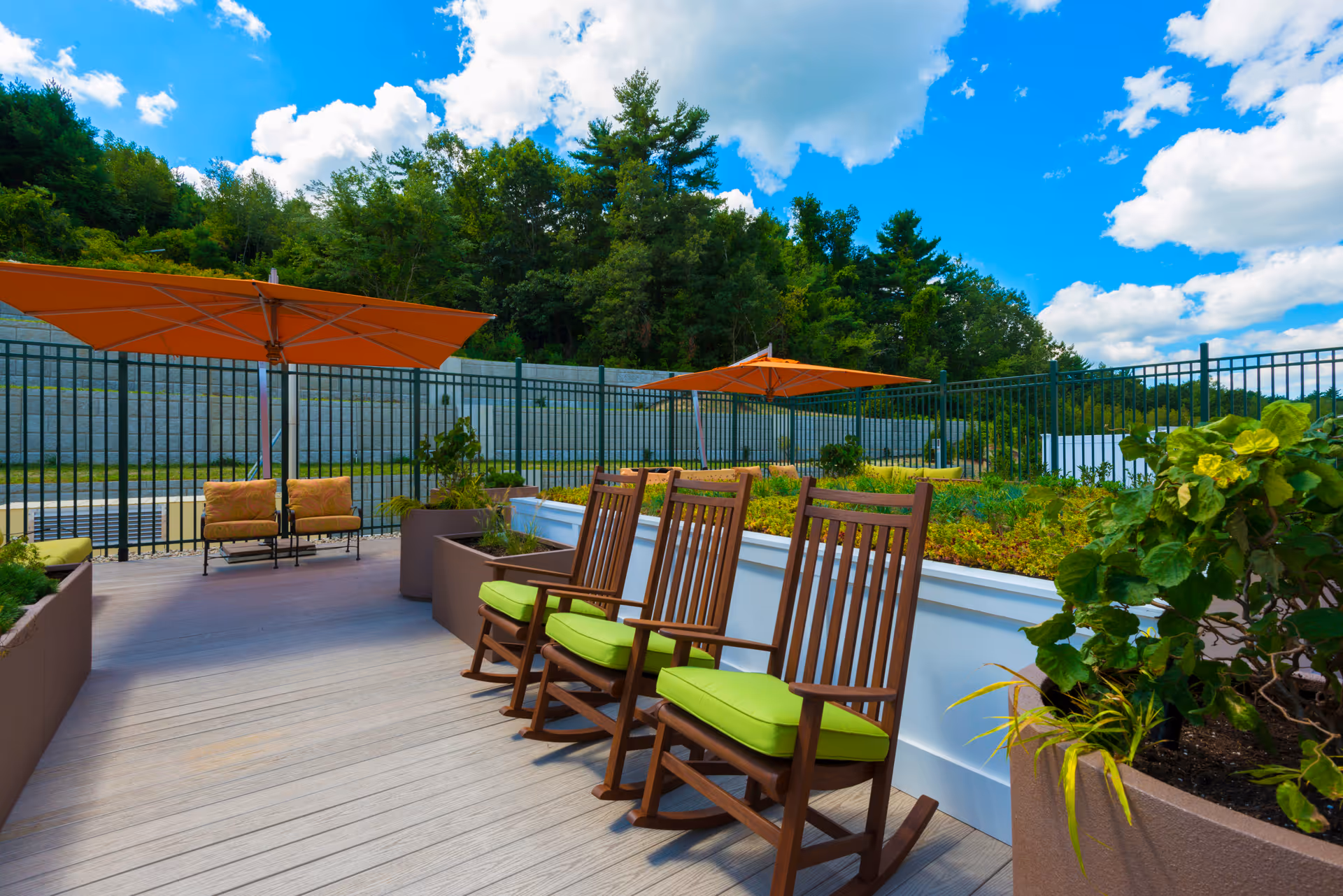 Outdoor patio area with wooden rocking chairs featuring green cushions, large orange umbrellas providing shade, potted plants, and a fenced garden area with greenery and trees in the background under a partly cloudy blue sky.