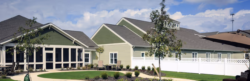 Exterior view of a senior living facility with green and beige buildings, a white fence, small trees, and a well-maintained lawn under a partly cloudy sky.