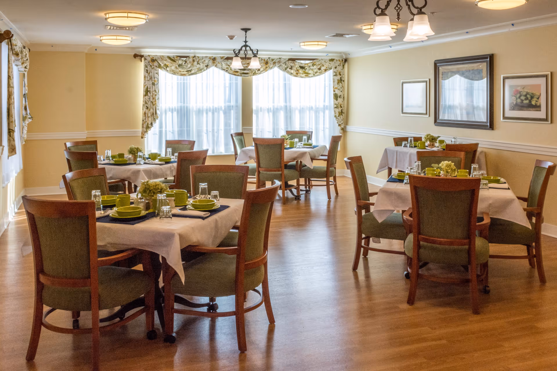 A bright dining room with several tables covered in white tablecloths, each set with green plates, cups, and glasses. The room has wooden chairs with green upholstery, large windows with floral curtains, framed artwork on the walls, and warm lighting fixtures on the ceiling.