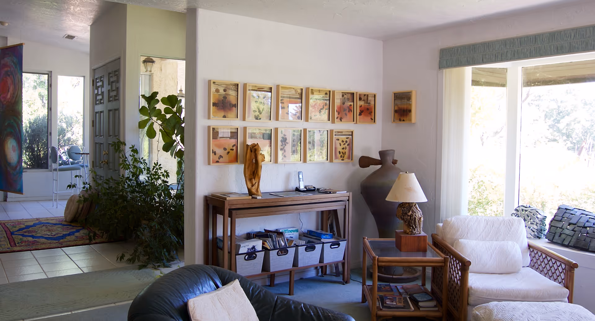 A bright and cozy living room area with a large window letting in natural light. The room features a white cushioned armchair, a wooden side table with a lamp, a console table with decorative items and storage bins underneath, and framed artwork on the wall. There are plants and a colorful rug visible in an adjacent room through an open doorway.