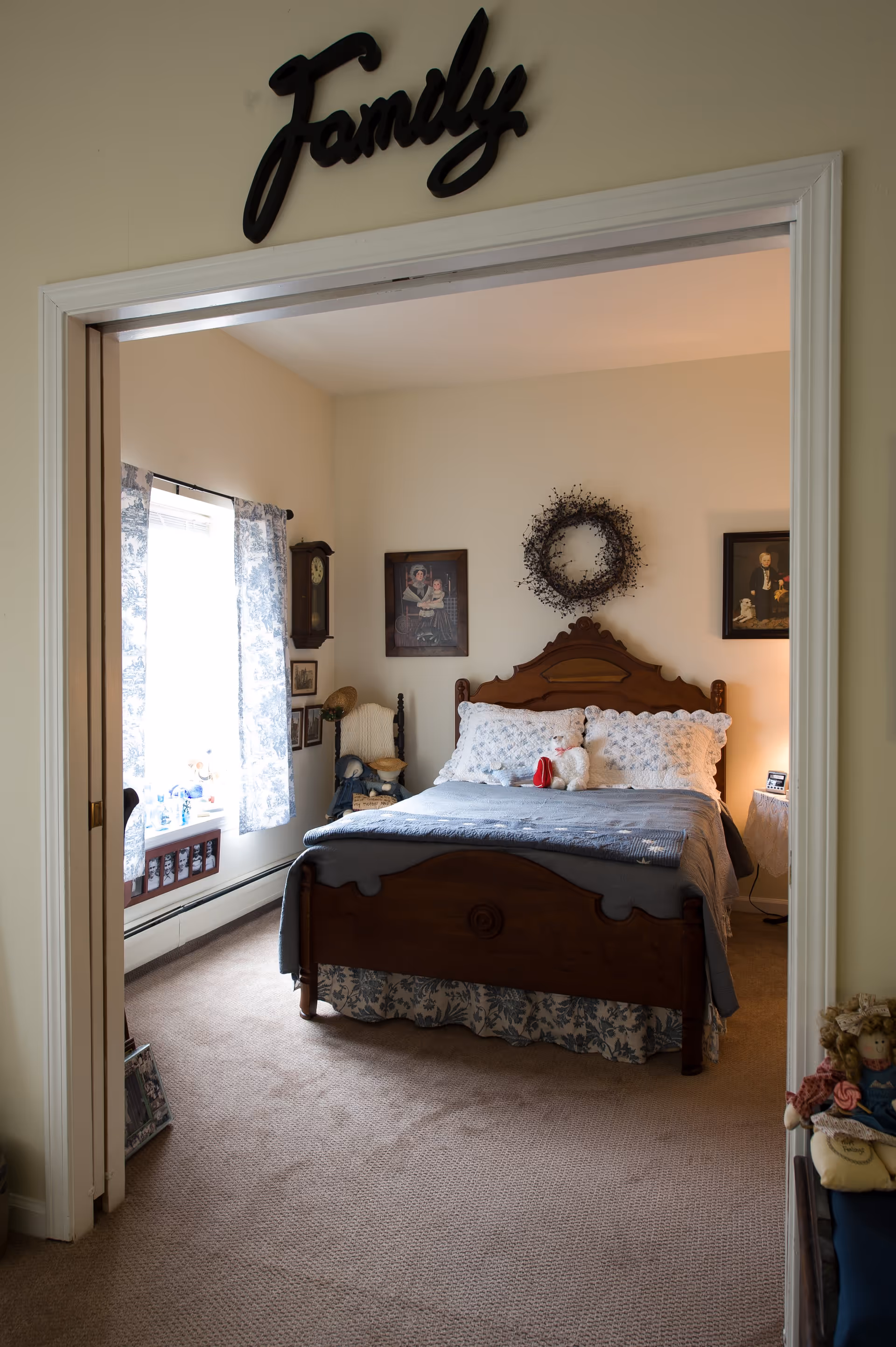 View into a cozy bedroom with a wooden bed frame, a blue and white bedspread, and decorative pillows. Above the bed hangs a wreath and two framed pictures on the wall. A window with blue patterned curtains lets in natural light. The room has beige carpet and a small side table with a lamp. The doorway has a black decorative sign above it that reads 'Family'.