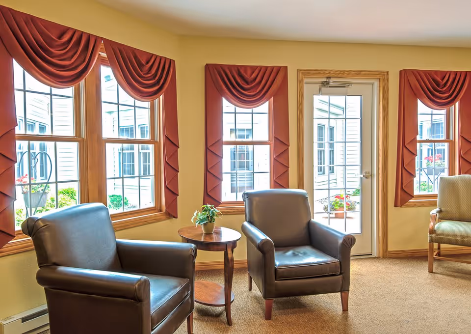 A cozy sitting area in a senior living facility with two dark leather armchairs and one upholstered armchair arranged around a small wooden round table with a potted plant. The room has beige walls, carpeted floor, and three large windows with red draped valances. A glass door with a wooden frame leads outside, showing a glimpse of a courtyard with flowers.