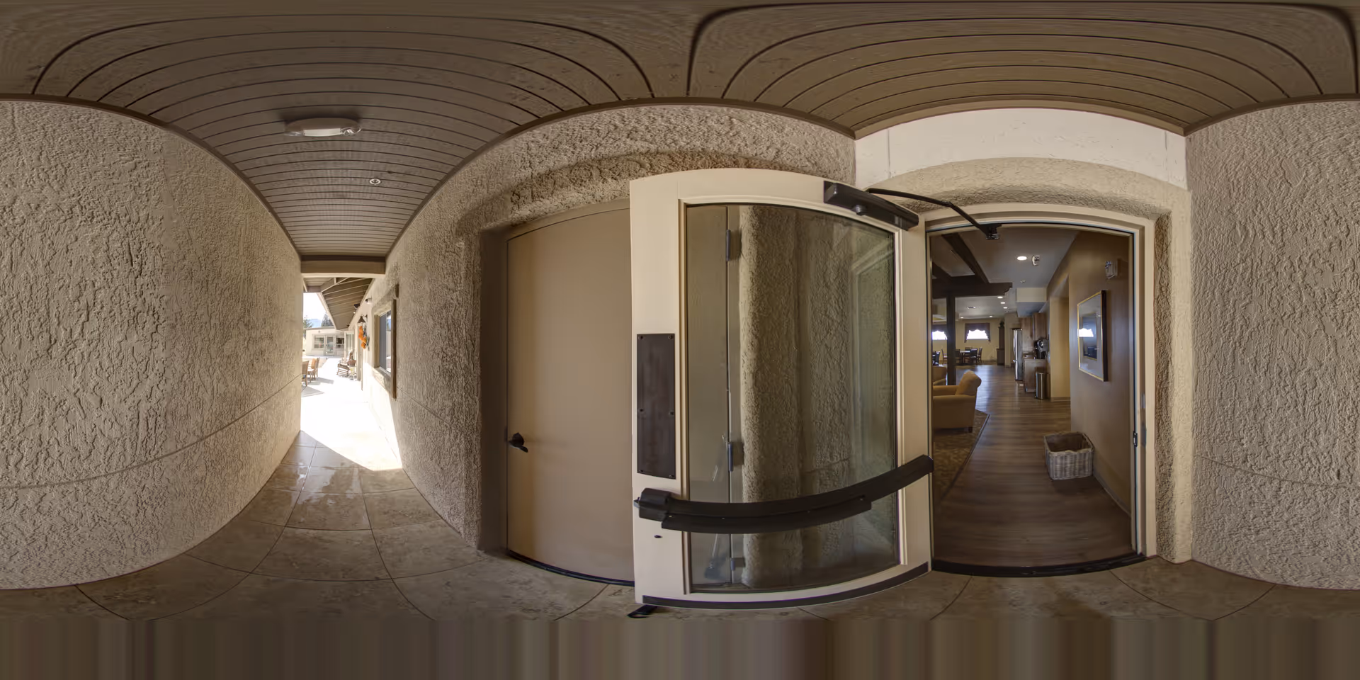 Open glass entry door leading from a shaded stucco exterior corridor into a warm interior common living area with seating.