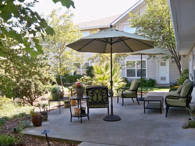 Outdoor patio area at Marbella Oroville with cushioned chairs, a table with potted plants, and a large umbrella providing shade. The patio is surrounded by greenery and trees, with a building visible in the background.