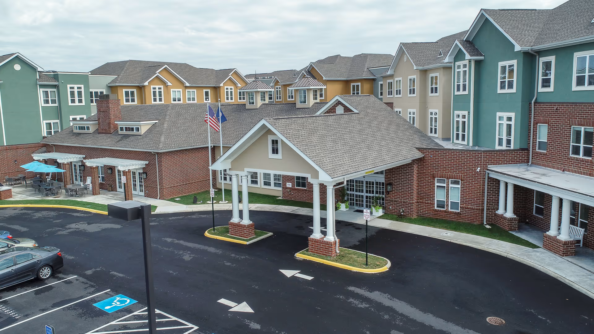 Exterior view of Harmony at Spring Hill senior living facility showing a multi-story building with green, beige, and yellow sections, a covered entrance with white columns, American and state flags, outdoor seating with umbrellas, and a parking lot with a handicap parking space.
