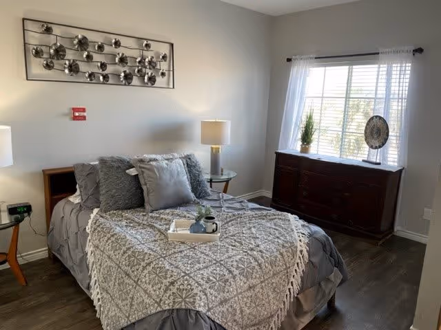 A cozy bedroom with a neatly made bed covered with a gray and white patterned blanket and multiple pillows. There is a tray with a small plant and a cup on the bed. The room has wooden flooring, a window with white curtains, a dark wooden dresser with decorative items on top, and two bedside tables with lamps. A modern metal wall art piece is mounted above the bed.